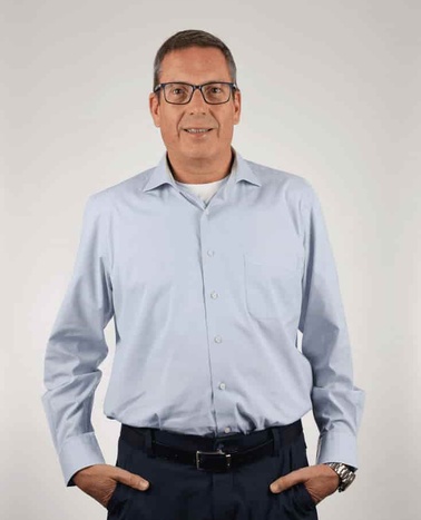 A man in a light blue shirt and black pants standing with his hands on his hips against a gray background.