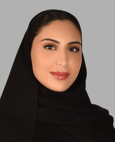 Portrait of a smiling woman wearing a black hijab against a gray background.