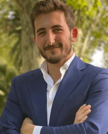 A young man in a blue blazer and white shirt smiling outdoors with trees in the background.