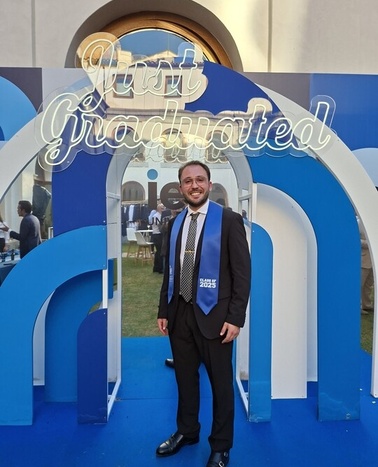 A man in a suit stands proudly in front of a graduation backdrop.