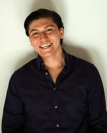 A young man in a dark blue shirt smiling at the camera against a light background.
