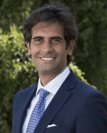 A man in a blue suit and tie smiling outdoors with greenery in the background.