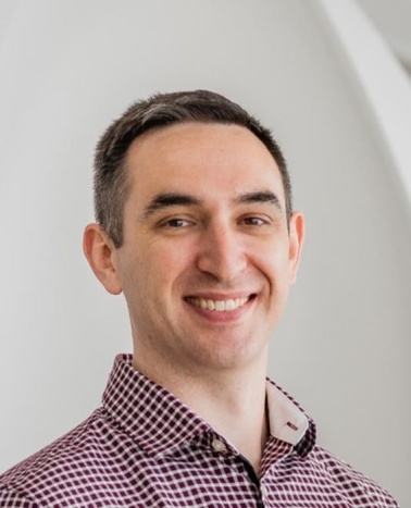 A smiling man in a red and white checked shirt standing against a light gray geometric background.