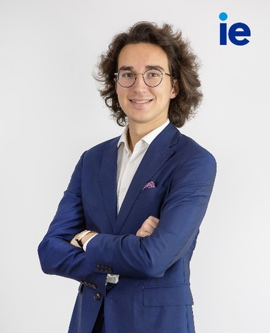 A young man in a blue suit with a white shirt and a pink pocket square stands confidently against a white background with the 'ie' logo above his shoulder.