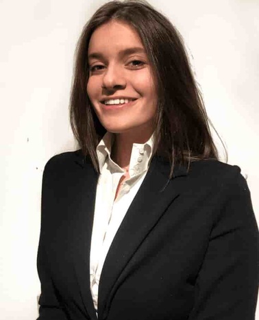 A young woman in a black blazer and white shirt smiling against a plain background.
