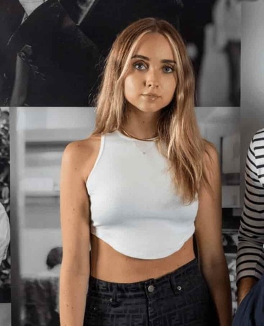A young woman with light brown hair wearing a white sleeveless top and black pants, standing against a black and white photo backdrop.