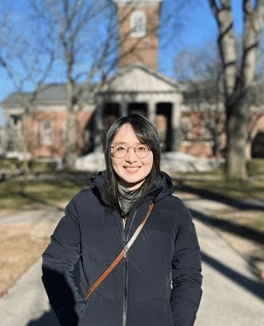 A woman smiling in front of a historic building with a clock tower on a sunny day.