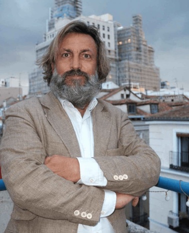 A man with a beard stands on a balcony with city buildings in the background.