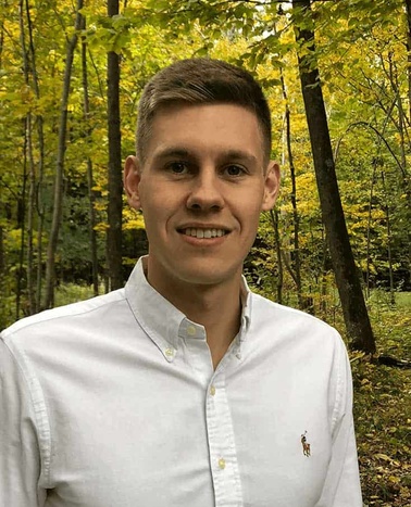 A young man in a white shirt smiling in a lush forest during autumn.