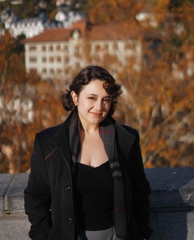 A woman smiling at the camera with an autumnal landscape and buildings in the background.