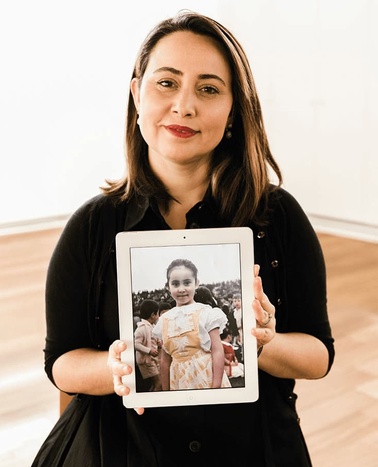 A woman in a black shirt holds a tablet displaying a black and white photo of a young girl.