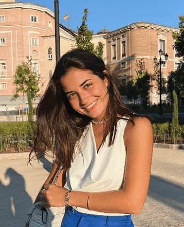 A young woman smiling in a sunny outdoor setting with a building in the background.