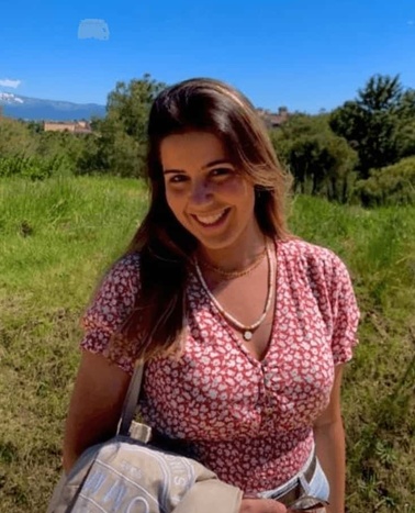 A smiling woman wearing a floral dress standing in a grassy field on a sunny day.