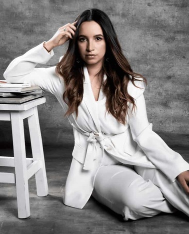 A woman in a white suit seated beside a white stool with books, posing thoughtfully in a studio setting.