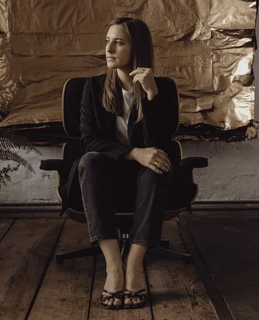 A woman in a thoughtful pose sits on a chair, dressed in a dark blazer and jeans, against a rustic backdrop.