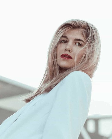 A young woman with blonde hair and red lipstick, looking over her shoulder with a thoughtful expression, set against a soft, blurred background.