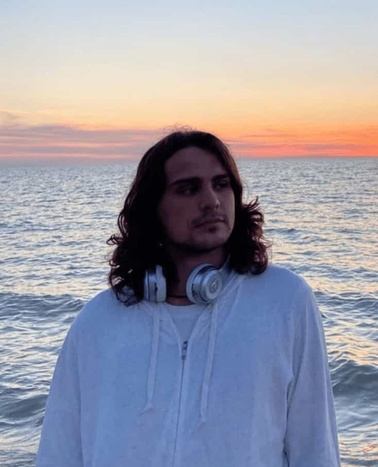 A young man with long hair and headphones around his neck stands before a sunset over the ocean.