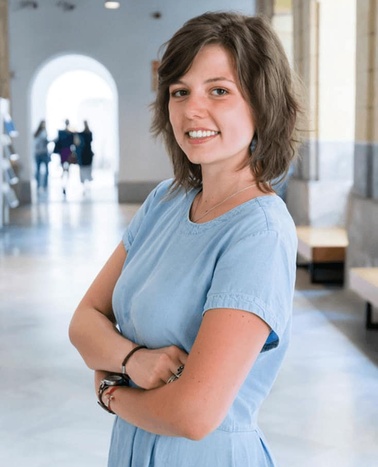 A young woman in a blue dress smiling at the camera in a hallway with people in the background.