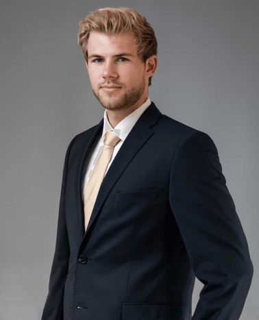 A young man in a dark suit and light shirt, looking confident with a slight smile.