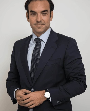 A confident man in a dark suit, light blue shirt, and dark tie, standing with his arms crossed against a plain background.