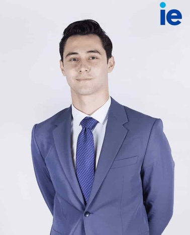 A man in a blue suit with a tie stands against a light background, with the logo 'ie' visible above his head.