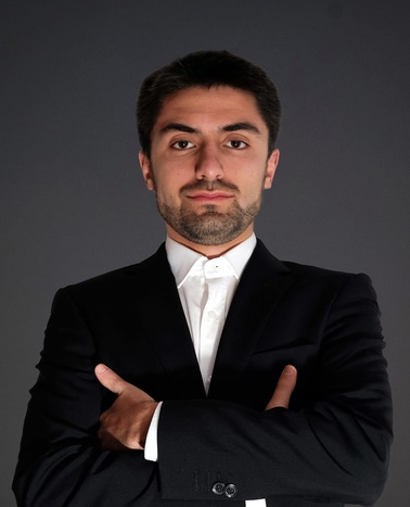A confident young man in a black suit and white shirt stands with his arms crossed, looking directly at the camera.