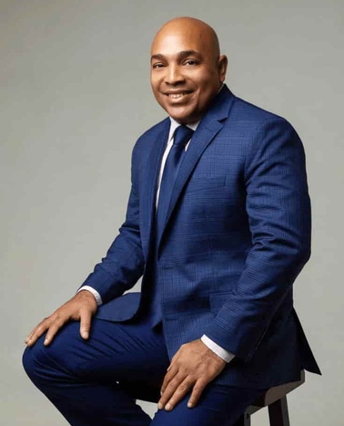 A man in a blue suit is smiling and sitting on a stool.