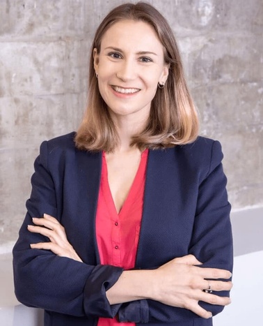 A professional woman in a blue blazer and red blouse smiling confidently with arms crossed in an office setting.