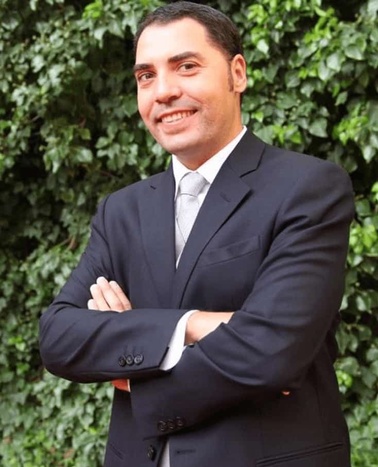 A smiling man in a dark suit standing with his arms crossed in front of a green leafy backdrop.
