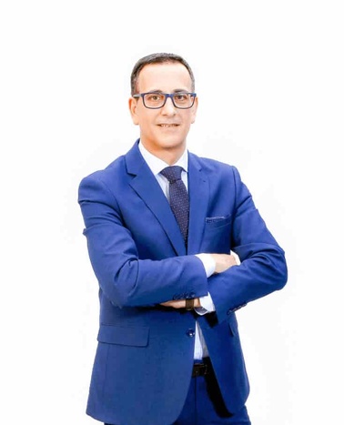 A man in a blue suit and glasses is standing with his arms crossed.