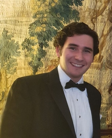 A young man in a tuxedo smiling in front of a decorative tapestry.