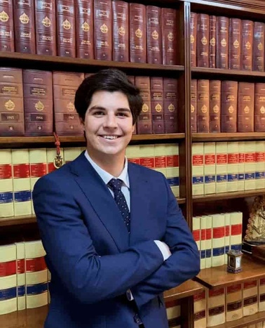 A young man in a blue suit and tie smiling in front of a bookshelf filled with red and gold bound books.