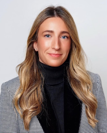 A professional portrait of a woman with long blonde hair wearing a houndstooth jacket and a black turtleneck.