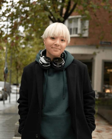 A young person with blonde hair smiling at the camera, wearing headphones around their neck and a dark coat over a teal hoodie, standing on a city street.