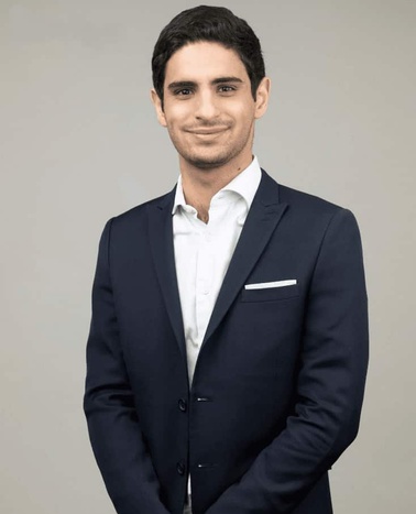A young man in a dark blue suit and white shirt, smiling at the camera against a gray background.