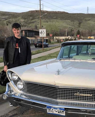 A person standing next to a classic car on a roadside with hills in the background.