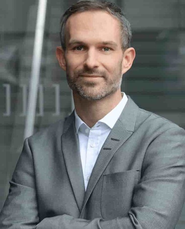 A confident man in a gray suit stands with his arms crossed in front of a glass building.