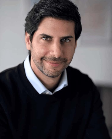 A portrait of a smiling man with dark hair, wearing a dark sweater and a white shirt.