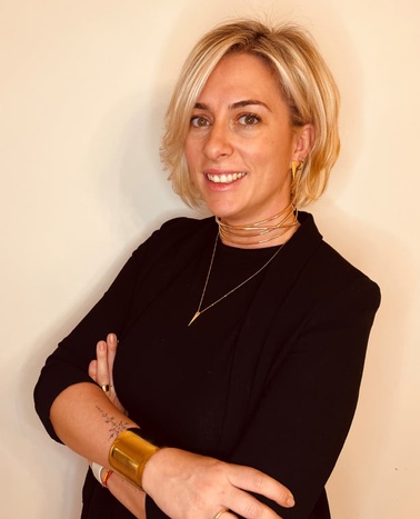 A confident woman with short blonde hair, wearing a black top and multiple gold necklaces and bracelet, stands against a cream-colored background.