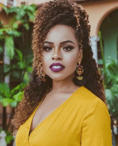 A portrait of a woman with curly hair wearing a yellow dress and bold makeup.