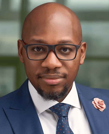 A professional portrait of a man in a blue suit, white shirt, and tie, wearing glasses.
