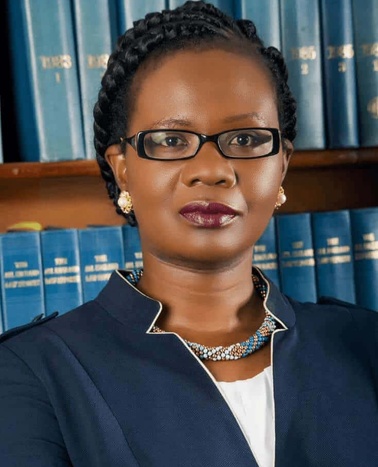 A professional portrait of a woman with braided hair, wearing glasses and a dark blue blazer, standing in front of a bookshelf filled with blue books.