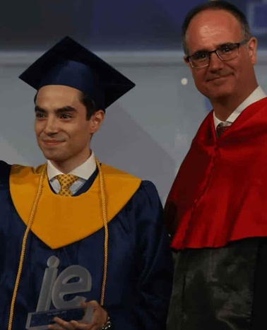 A young graduate in a blue and yellow cap and gown receives an award from an older man in a red and black academic robe.