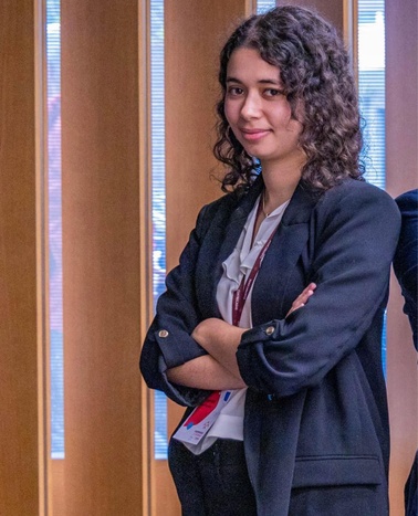 A young woman in a blazer and ID badge stands with crossed arms, smiling in an office environment.