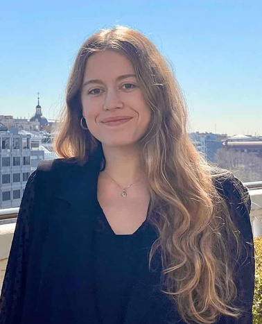 A young woman with long blonde hair, smiling, dressed in a black outfit, standing on a balcony with a cityscape in the background.