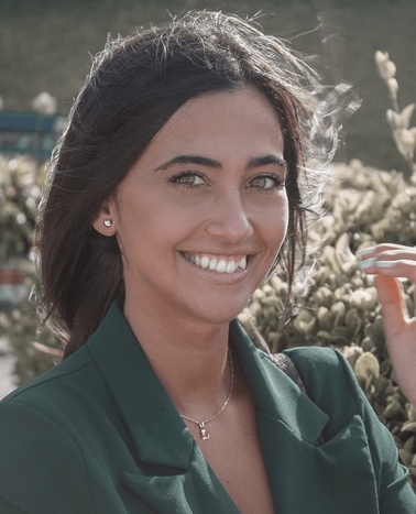 A smiling young woman in a green blouse standing outdoors with sunlight highlighting her hair.