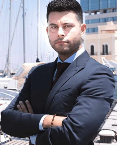 A confident man in a business suit stands with his arms crossed at a marina.