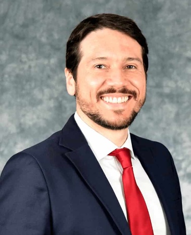 A smiling man in a blue suit and red tie standing against a grey textured background.