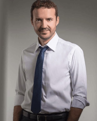 A professional portrait of a man in a white shirt and blue tie, standing against a grey background.