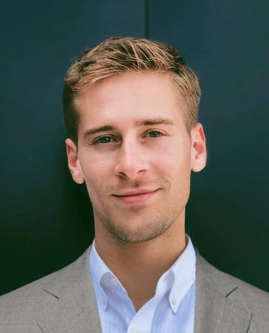 A portrait of a young man with short hair, wearing a grey blazer and a blue shirt, set against a dark blue background.